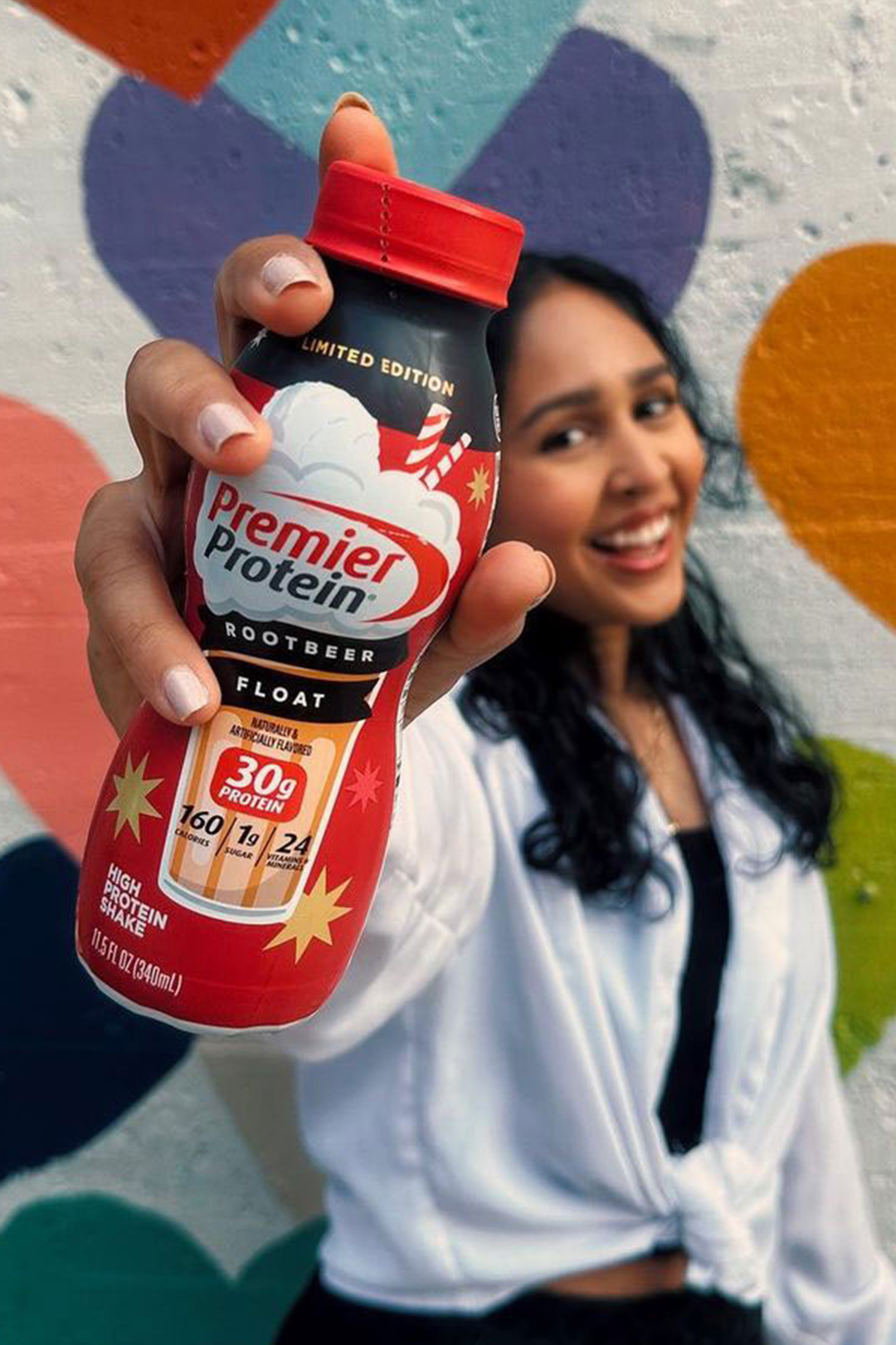 Woman in front of a mural holding up a bottle of Premier Protein Root Beer Float