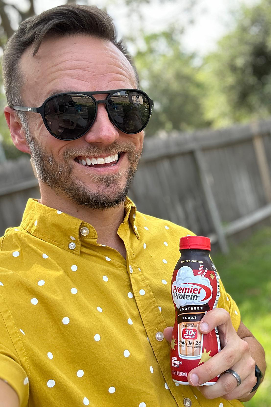 Man in sunglasses smiling outdoors while holding a Premier Protein Root Beer Float protein shake